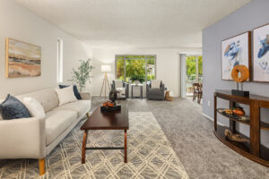 Apartment living room with neutral décor, carpet flooring, large windows, and sliding door to a private balcony at Wildwood Apartment Homes in Issaquah, Washington.